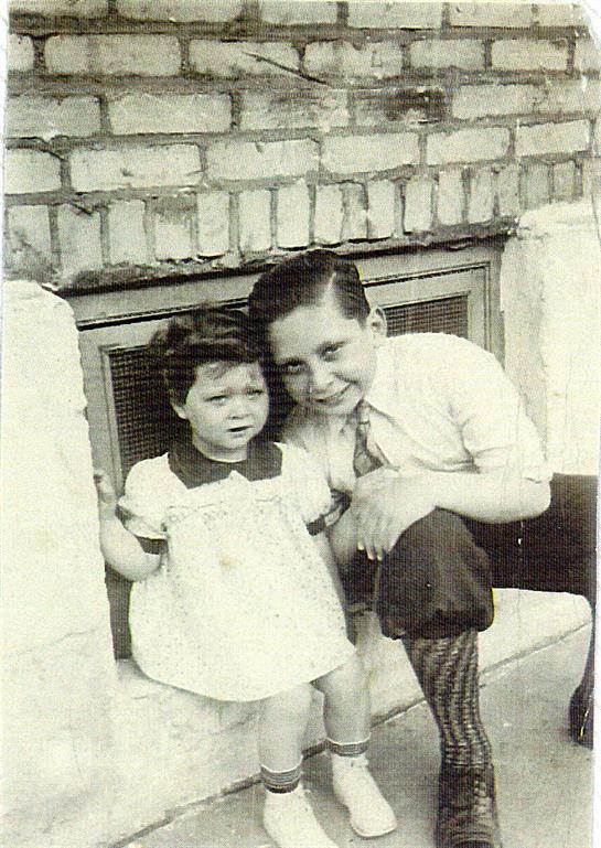 Two children stand together by a brick wall, smiling and sharing a moment of camaraderie.