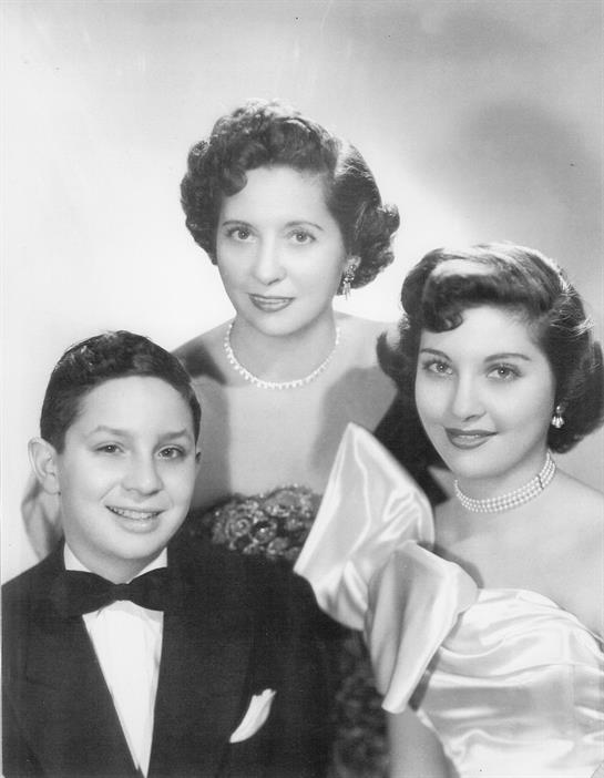 Three individuals pose in formal wear with elegant hairstyles in a classic setting.
