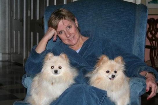 A woman wearing a blue robe smiles while sitting on a sofa with two fluffy dogs by her side.