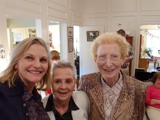 Three women, two older and one younger, share smiles in a cozy, memory-filled setting.