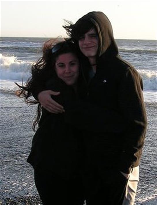 A couple stands together on the beach, wrapped in each other's arms as waves crash nearby.
