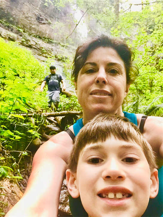Two family members pose for a selfie while hiking through vibrant greenery with a waterfall nearby.