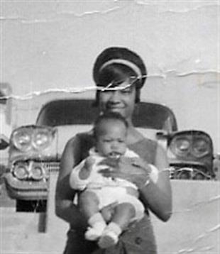 A woman smiles while holding her baby in front of a vintage car on a sunny day.