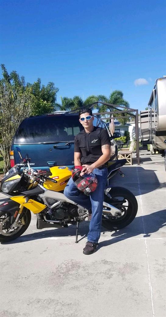 A young man in sunglasses sits on a yellow motorcycle by parked cars under a clear sky.