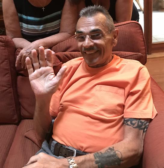 A man dressed in an orange shirt smiles and waves while sitting comfortably on a couch.