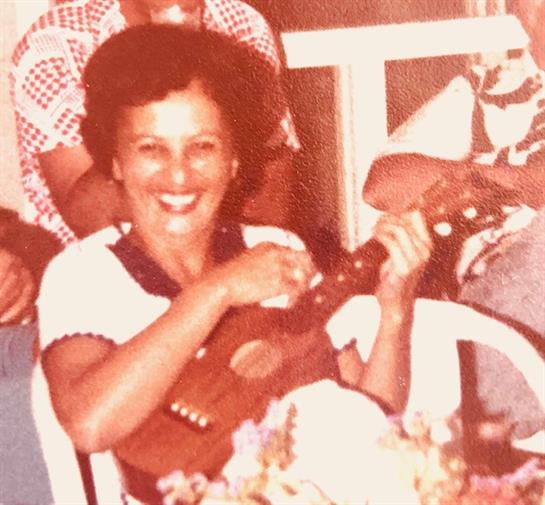 Woman joyfully strums a ukulele, surrounded by friends in a lively setting.