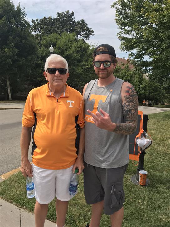 Two men stand together, one in an orange shirt and the other in a tank top, both smiling.