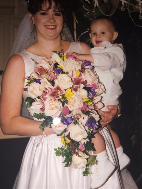 A woman in a white dress smiles while holding a colorful bouquet and a happy child at a wedding.