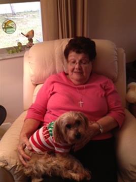 An elderly woman relaxes in a comfortable chair, holding a small dog in holiday attire.