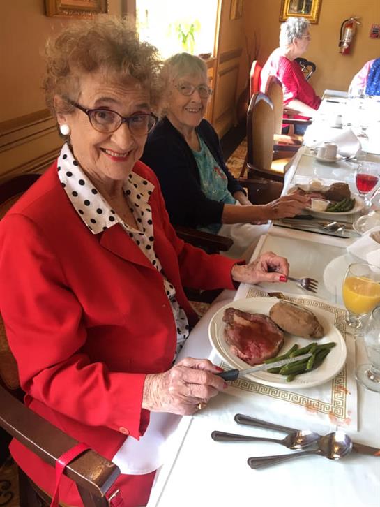 An elderly woman with glasses smiles while holding a fork next to her dish of meat and vegetables.