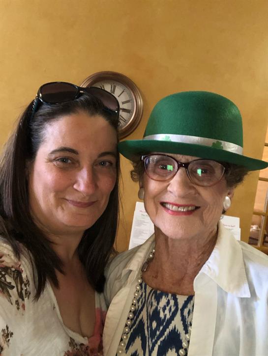 Two women pose happily in a cheerful, sunlit space with yellow walls and a vintage clock.