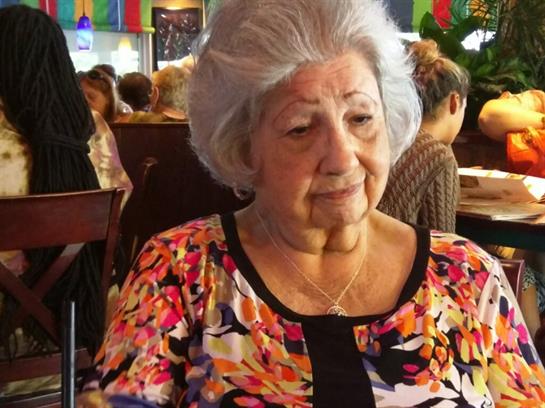 A cheerful elderly woman sits at a table in a busy restaurant, surrounded by friends.