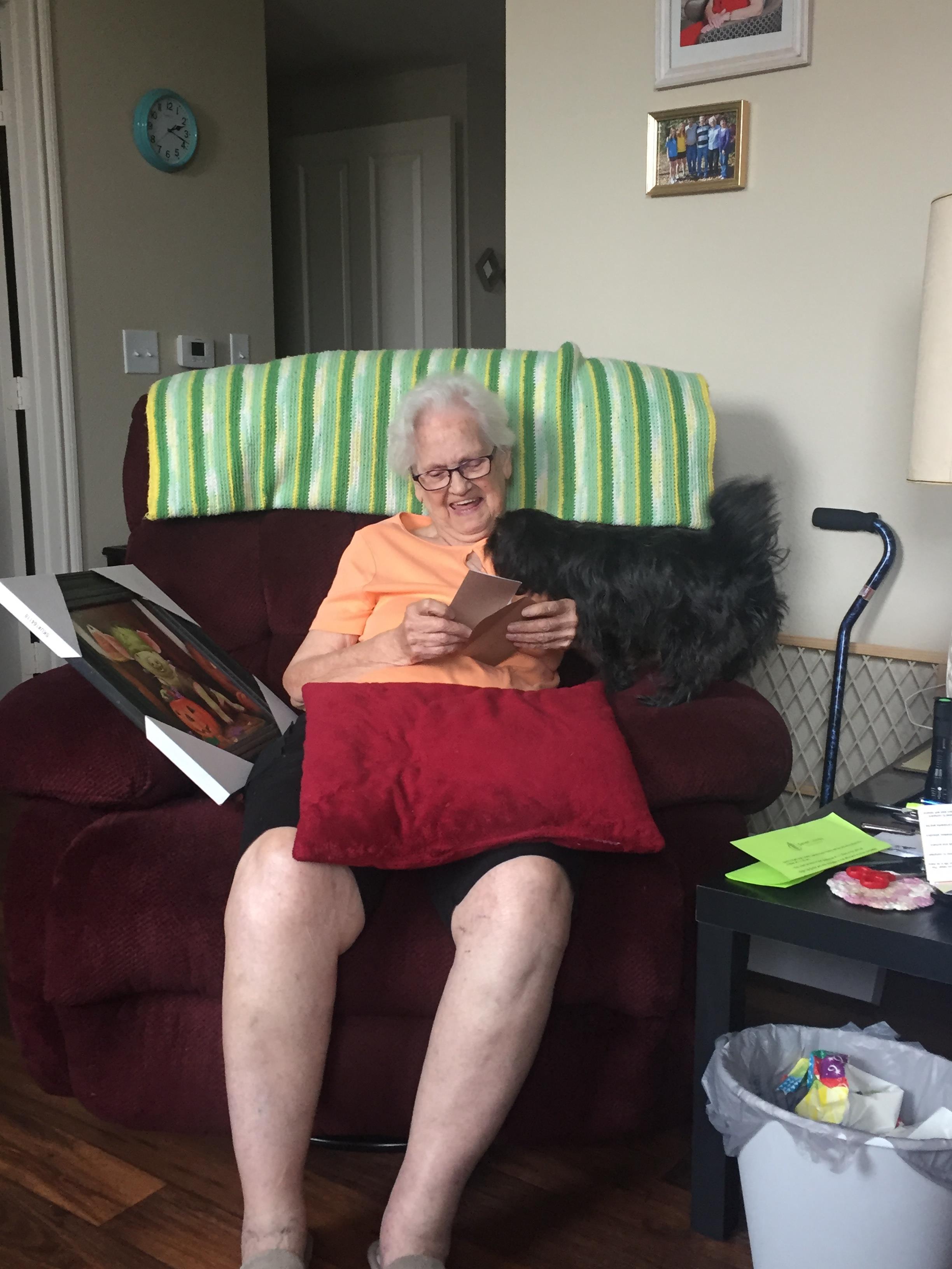 Smiling elderly woman enjoys a card as her dog leans in for affection in a warm living space.