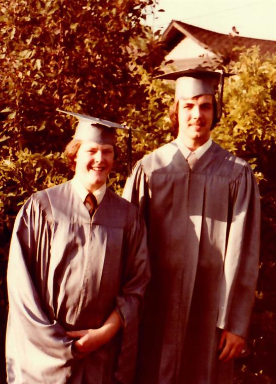 Two graduates proudly pose in caps and gowns amidst vibrant greenery on a sunny day.