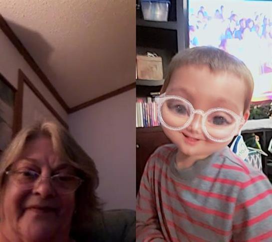 A grandmother smiles warmly as her playful grandchild poses with oversized glasses at home.