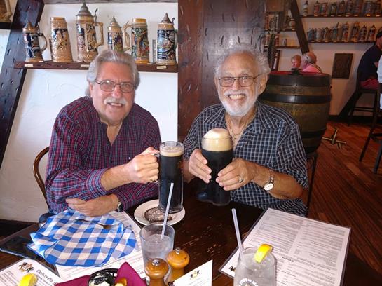 Two friends enjoy their lunch, smiling and toasting with large glasses of dark beer in a restaurant.