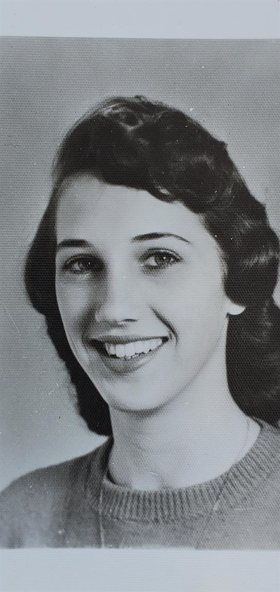 A cheerful young woman poses with a bright smile, showcasing her wavy hair and classic style.