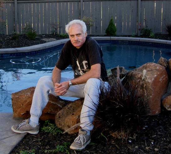 A senior man relaxes on a rock by a pool in a landscaped backyard during the evening.