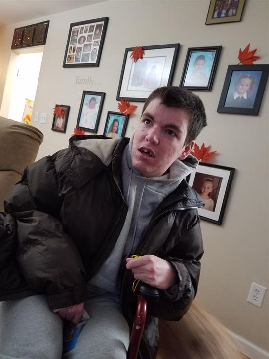 A young man sits quietly in a chair looking around a warmly decorated room with family pictures.