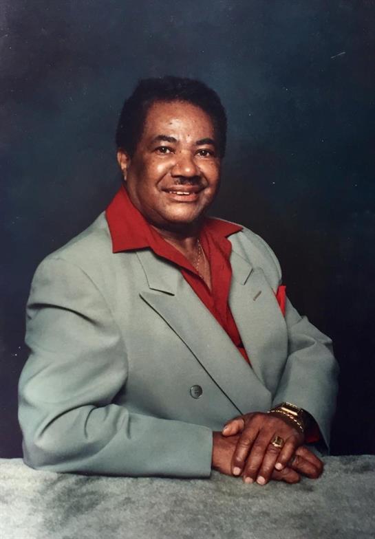 A man poses with a warm smile, wearing a light-colored suit along with a vibrant red collar.