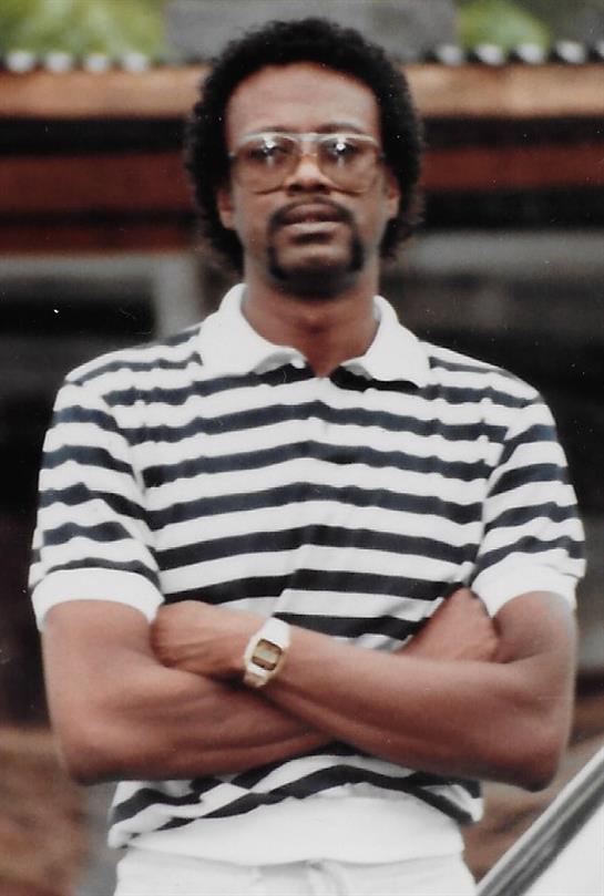 A man with glasses stands confidently in a striped polo shirt, arms crossed, under clear sky.