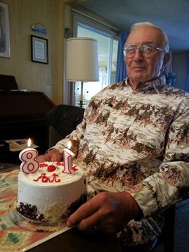 An elderly man sits with a birthday cake marked with 81, smiling as he celebrates his special day.