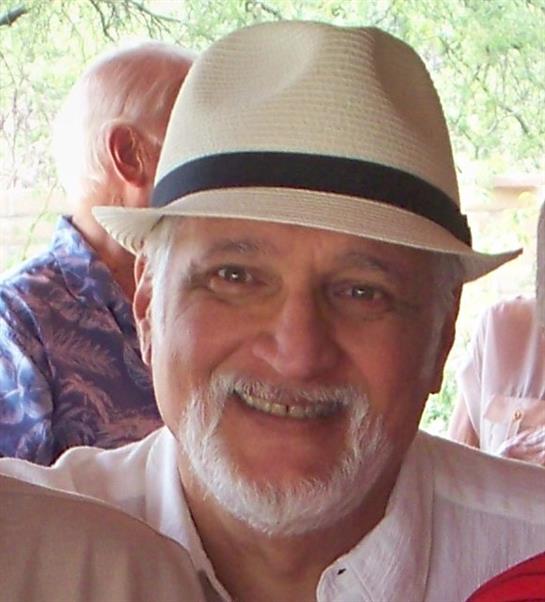 A joyful man wearing a white fedora is smiling at a friendly gathering outdoors with greenery.