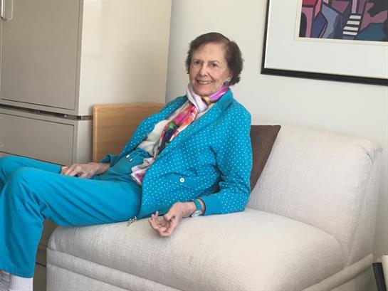 A woman wearing a bright blue suit smiles while seated on a stylish sofa in a warm room.