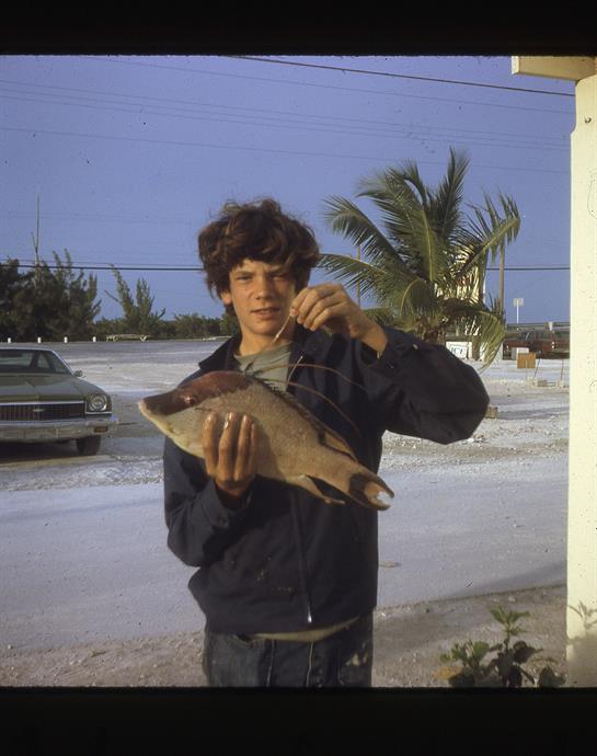 Teenager holds a large fish in a coastal area with palm trees and a classic car nearby.