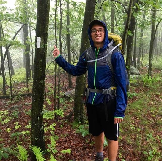 Hiker stands under light rain, showcasing trail marker while exploring lush forest.