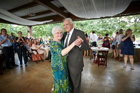 An elderly couple shares a romantic dance at a wedding reception surrounded by guests.