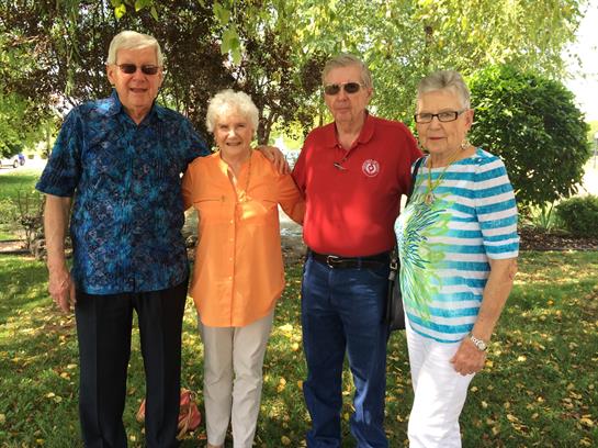 Four seniors stand together in a garden, smiling brightly on a sunny day.