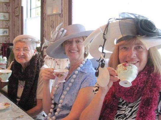 Three women smile and sip tea while wearing stylish hats at a cheerful gathering.