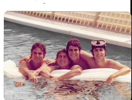 Group of four friends laughing and relaxing on inflatable rafts in a sunny swimming pool.