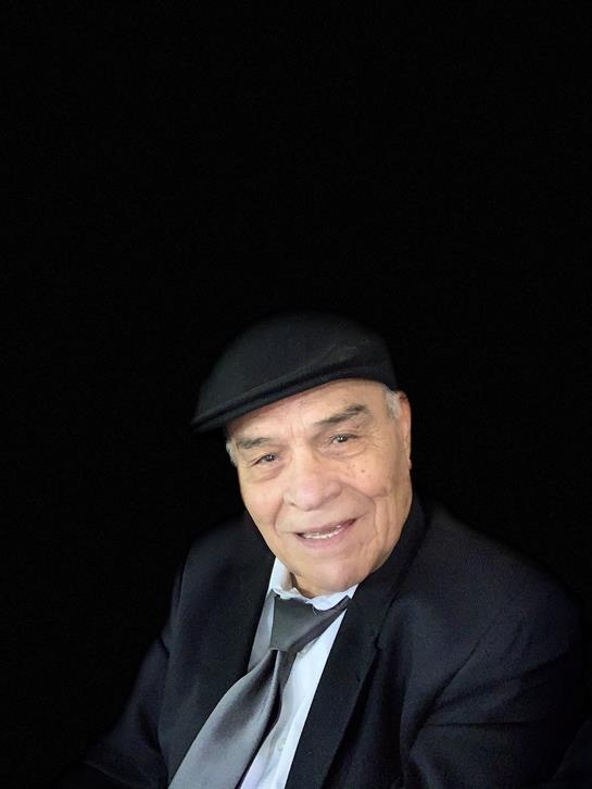 An elderly man dressed in a suit and cap smiles cheerfully, radiating warmth and charm indoors.