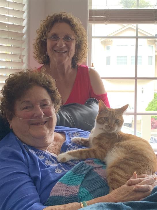 Two smiling women sit together on a couch with a cat, enjoying their time in a comfortable space.