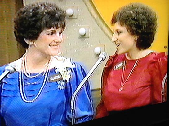 Two women smile and engage in conversation on stage during a festive game show event.