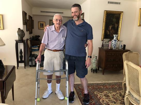 An elderly man using a walker stands beside a smiling caregiver in a warm household environment.
