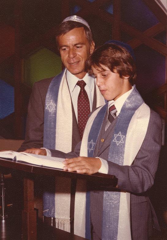 A young boy stands at a podium reading from a ceremonial book while an adult watches proudly.