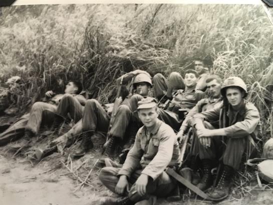 Group of soldiers relaxing among tall grass during a military mission in a forested environment.