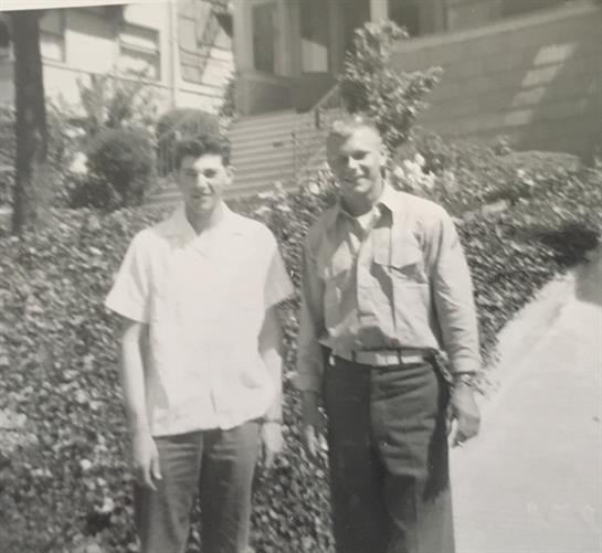 Two men stand together smiling in front of a house framed by bushes and trees.