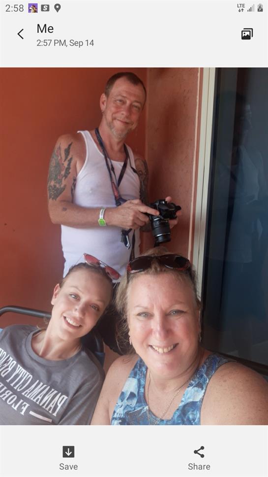 Three friends smile happily on a balcony, one holding a camera, enjoying a warm day together.