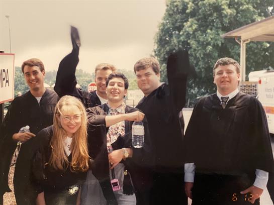 Six friends celebrate graduation outdoors, radiating joy in their outfits.