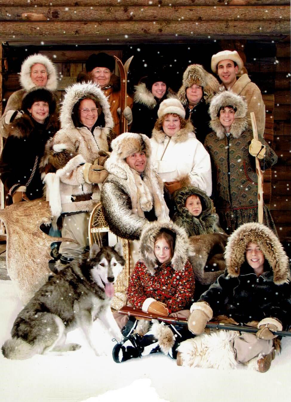 A large group dressed in warm, fur-lined clothing gathers in snowy scenery, surrounded by sled dogs.