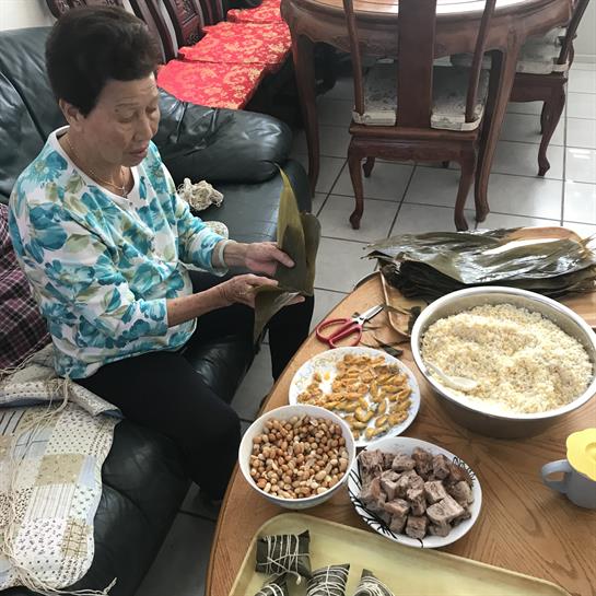 An elderly woman sorts ingredients for a traditional meal while sitting comfortably in her home.