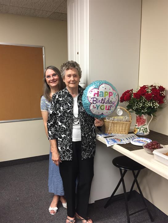 Two women celebrate a birthday, one holding a balloon, surrounded by flowers and treats.