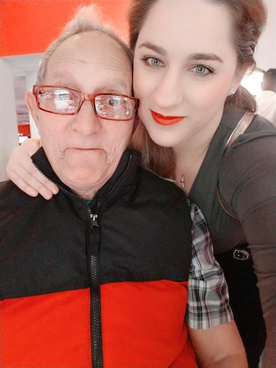 A young woman and an elderly man pose closely, smiling and enjoying their time indoors.