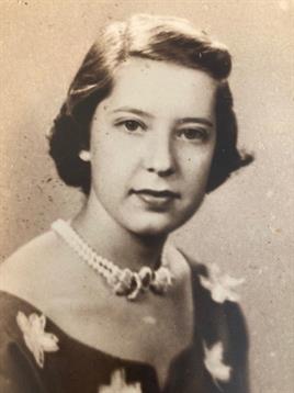 A young woman poses elegantly, wearing a vintage dress adorned with floral details and pearls.