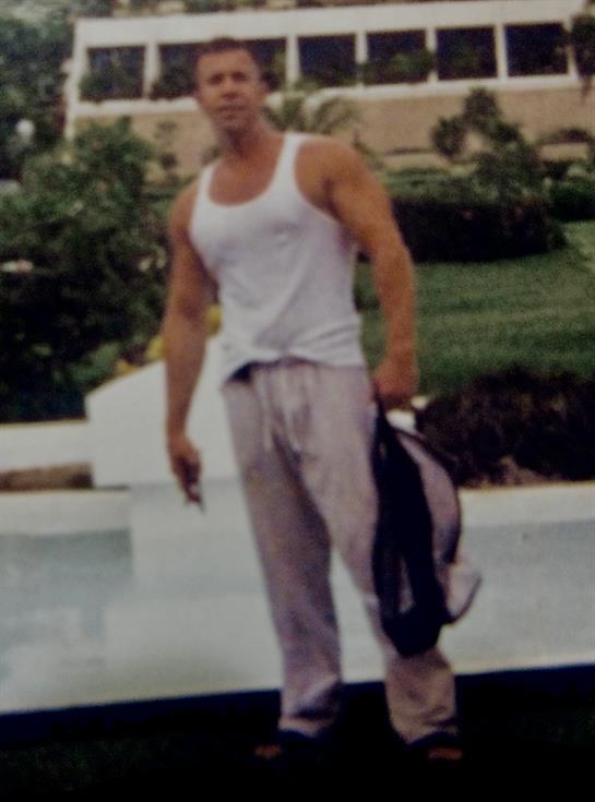 Man in tank top and sweatpants stands by a pool, holding a jacket, smiling in warm weather.