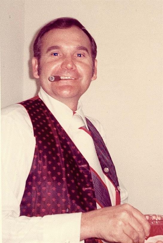 A mature man stands confidently, wearing formal attire with a cigar in his mouth, smiling indoors.